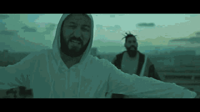 a man with a beard wearing a white hoodie stands next to another man with dreadlocks .