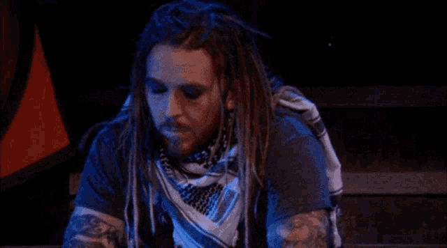 a man with dreadlocks and a scarf around his neck is sitting in a dark room with his eyes closed .