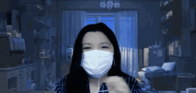 a woman wearing a white face mask looks at the camera in a dark room