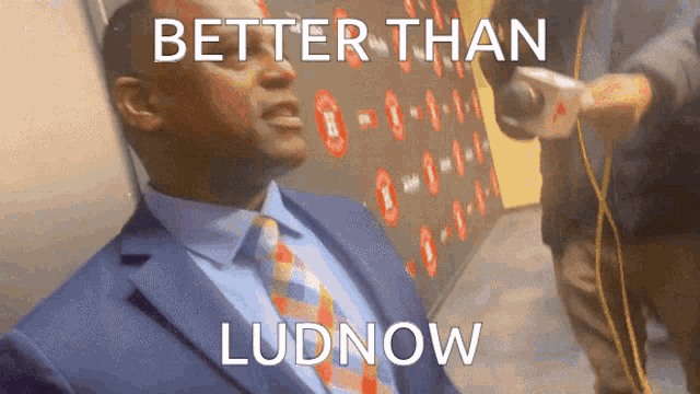 a man in a suit and tie is sitting in front of a wall that says " better than ludnow "
