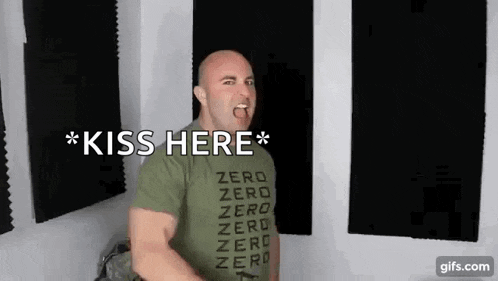 a man wearing a green shirt that says `` kiss here '' is standing in a room with his tongue out .