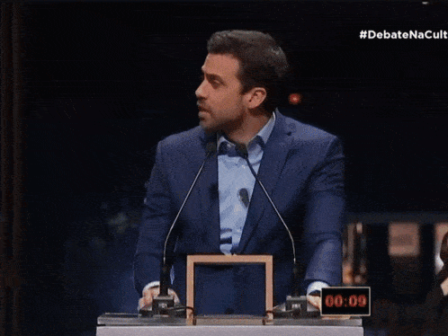 a man in a suit and tie stands in front of two microphones with the hashtag #debatenacult on the bottom right