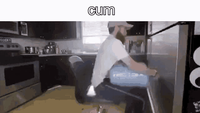 a man is sitting in a chair in a kitchen with the word cum on the bottom of the screen .