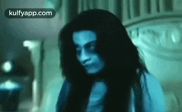 a woman with long hair and blue eyes is sitting on a bed in a room .