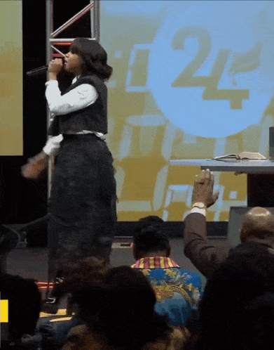 a woman sings into a microphone in front of a screen that says t24 live