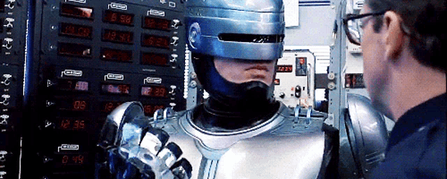a man in a helmet stands next to a robot that says ' robocop ' on the front of it