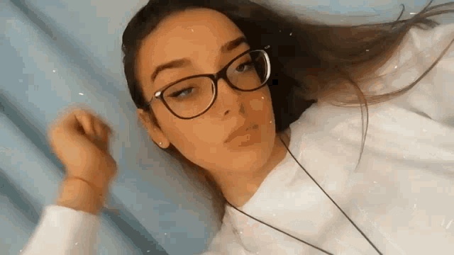 a young woman wearing glasses and a white sweater is laying down on a bed .