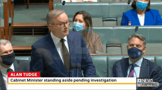 a man in a suit and tie is giving a speech in front of a microphone with the words alan tudge on the screen
