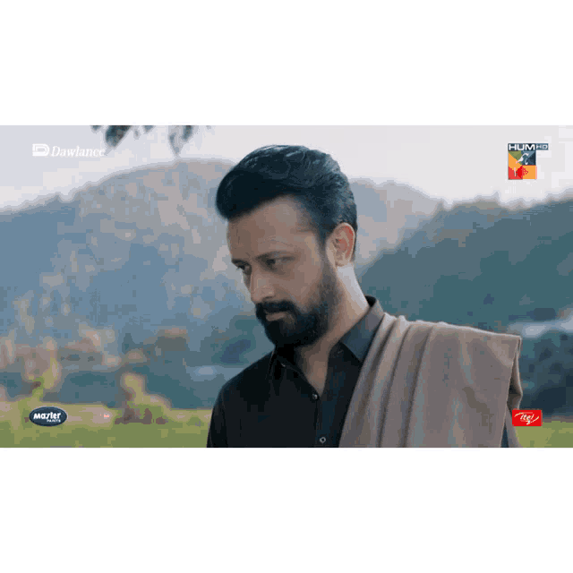a man with a beard is standing in a field with mountains in the background and a advertisement for itel on the bottom right