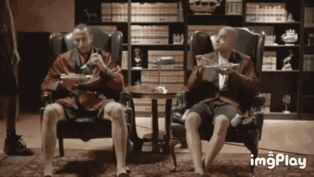 two men are sitting in chairs eating food with imgplay written on the bottom right