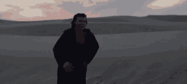 a man in a black coat is standing in the middle of a desert