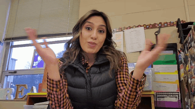 a woman wearing a plaid shirt and a black vest stands in front of a bulletin board with the letter f on it