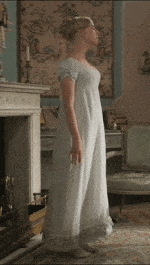 a woman in a white dress stands in front of a fireplace in a room