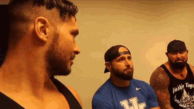 a man wearing a blue shirt with the letter h on it talks to two other men