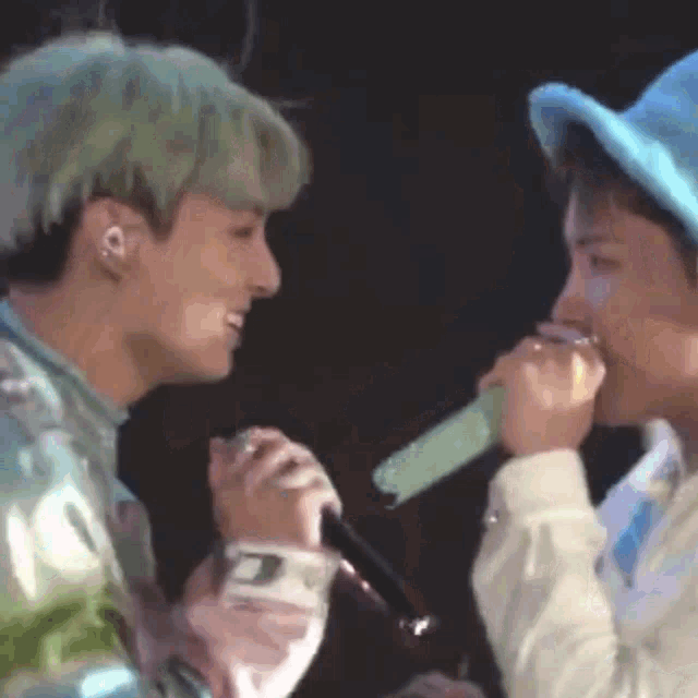 two men are singing into microphones on a stage and one of them is wearing a blue hat .