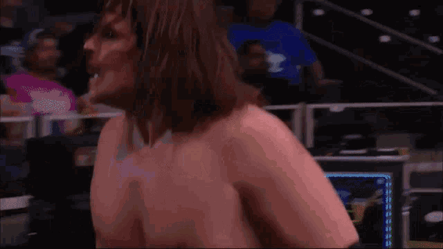 a shirtless wrestler with long hair stands in front of a crowd with a security guard in the background