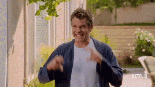 a man in a blue cardigan is standing in front of a house and making a funny face .