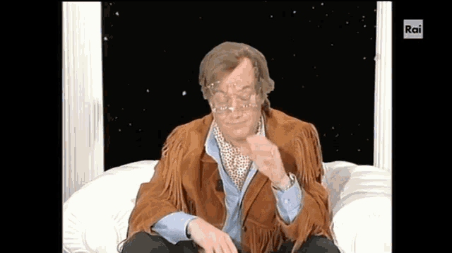 a man in a brown jacket is sitting on a white couch with rai written on the bottom right