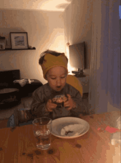a little boy wearing a yellow headband is sitting at a table with a bowl of food and a glass of water