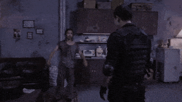 two men are standing in a living room and one of them is wearing a black vest that says ' army ' on it