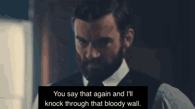 a man with a beard is standing in a dark room and talking to another man .