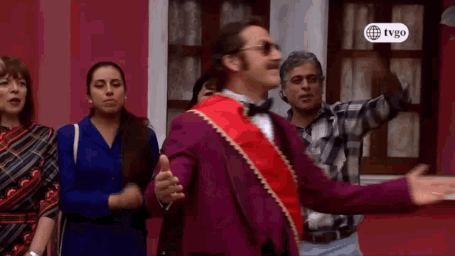 a man in a purple suit with a red sash is dancing in front of a tvgo sign