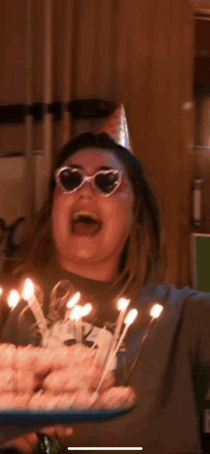 a woman wearing sunglasses and a party hat blows out candles on her birthday cake