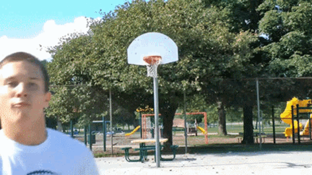 a man in a white shirt stands in front of a basketball hoop in a park