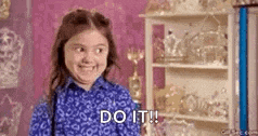 a little girl in a blue shirt is smiling and saying `` do it ! ''