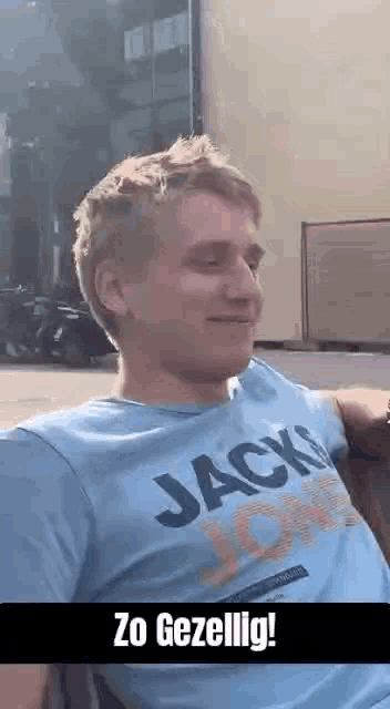 a man wearing a blue jack jones t-shirt is sitting on a bench and smiling .