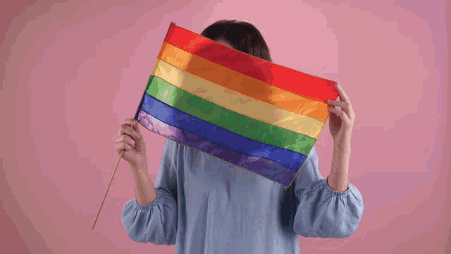 a woman wearing glasses holds a rainbow flag in front of her face