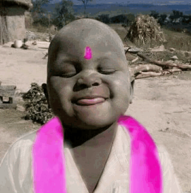a young boy with a pink ribbon around his neck and a pink bindi on his forehead is smiling .