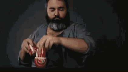 a man with a beard is sitting at a table holding two cups of coffee .