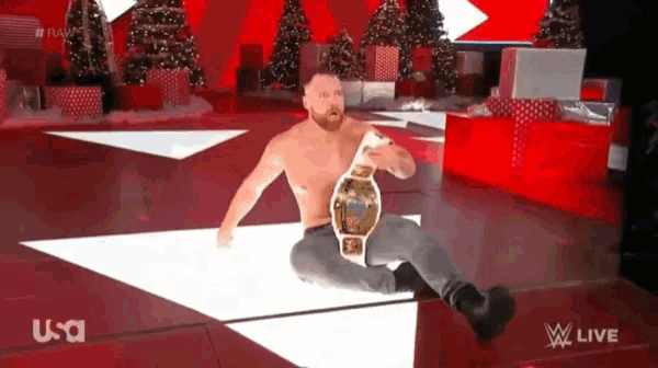 a shirtless wrestler is sitting on the floor holding a championship belt