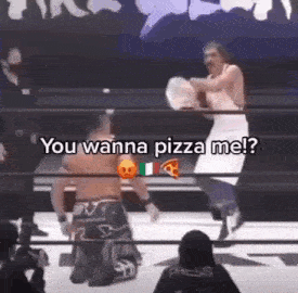 a man in an apron is kneeling down in a wrestling ring while another man throws pizza at him .
