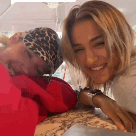two women are sitting at a table and smiling at the camera . one of the women is wearing a leopard print bandana .