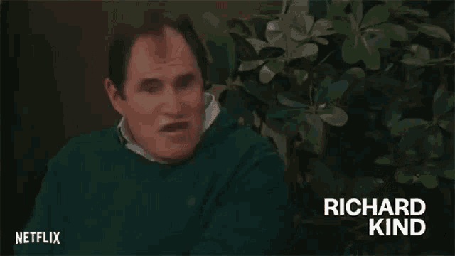 a man in a green sweater is sitting in front of a tree and making a funny face .