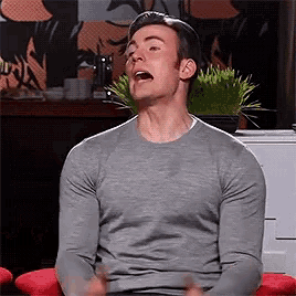 a man in a grey sweater is sitting on a red chair with his mouth open .