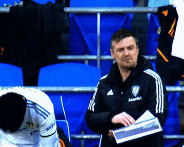 a man wearing a black adidas jacket stands in a stadium