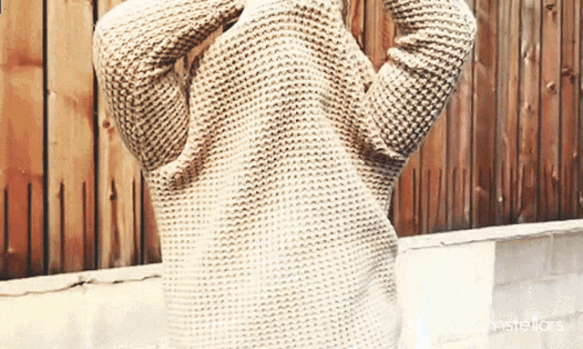 a person wearing a knitted sweater is standing in front of a wooden fence that says accumstellars