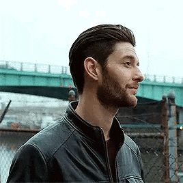 a man with a beard is wearing a black leather jacket and standing in front of a bridge .