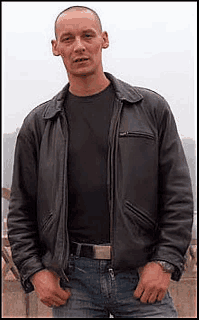 a man wearing a black leather jacket and jeans is standing in front of a building .