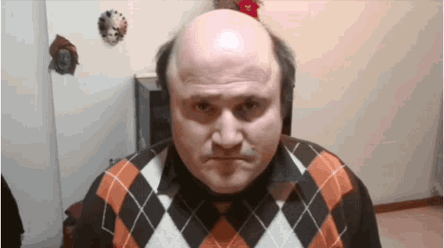 a bald man wearing a plaid sweater makes a funny face