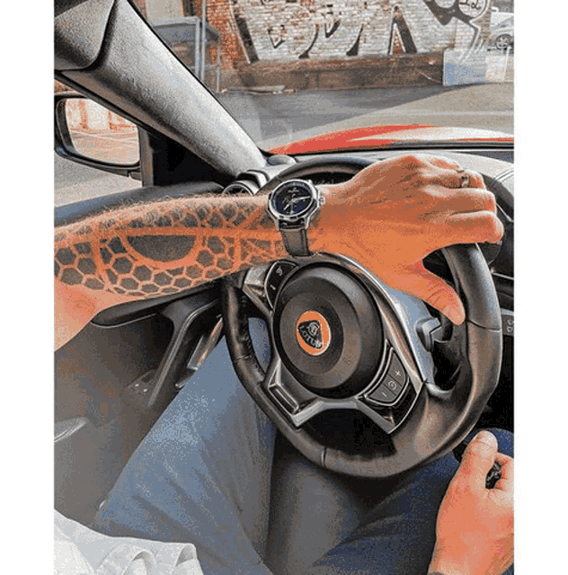 a man with a tattoo on his arm is driving a red car with a lotus logo on the steering wheel