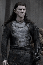 a man with long hair and armor is standing in front of a building .