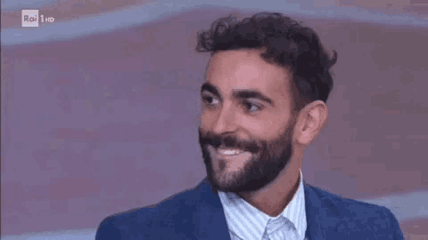 a man with a beard and curly hair is smiling and wearing a blue suit and striped shirt .