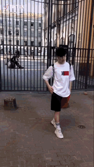 a young man wearing a white tommy hilfiger shirt and black shorts is standing in front of a fence .