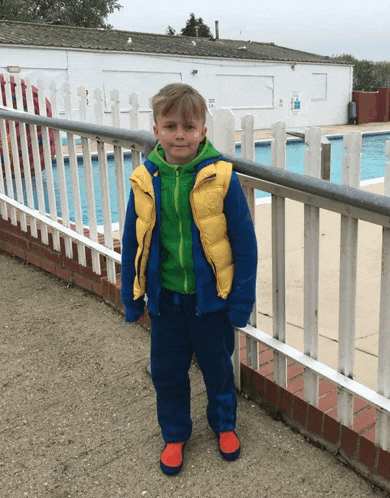 a young boy wearing a yellow vest and blue pants stands in front of a white fence