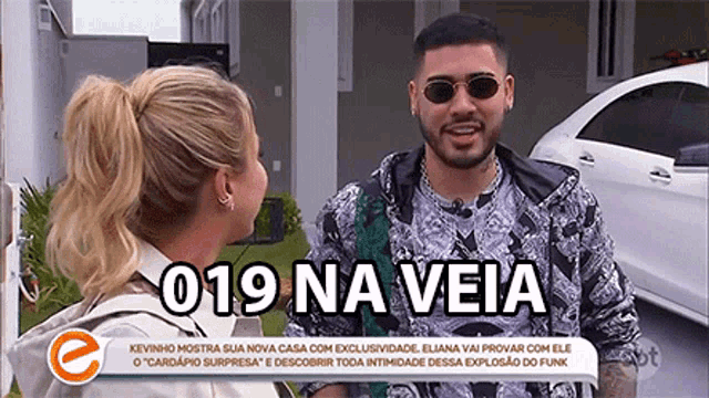 a man and a woman are talking in front of a car with the words 019 na veia on the bottom