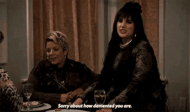 a woman is sitting at a table with a glass of wine and talking to a group of women .